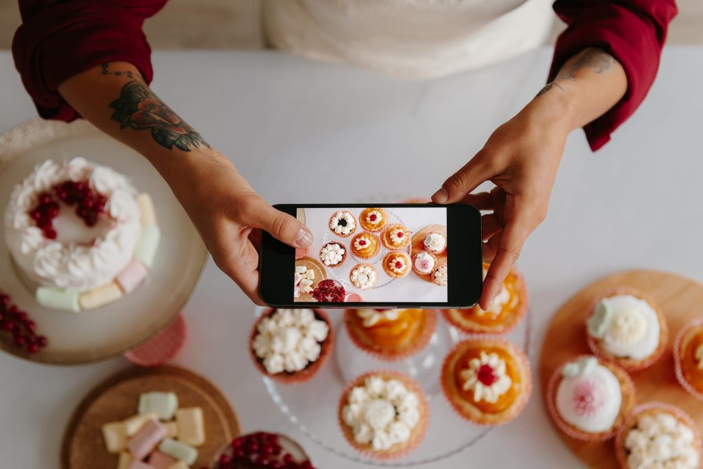 Top view of female baker using smart phone while photographing sweet food laying on the table